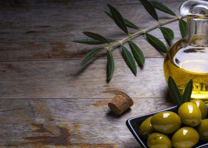 Primo piano di olive verdi appena raccolte accanto a una bottiglia di olio EVO dorato, grassi food trend